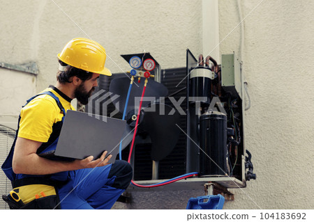 Licensed technician commissioned for outside hvac system annual maintenance, looking for refrigerant leaks. Certified expert doing air conditioner inspection, imputing data on laptop 104183692