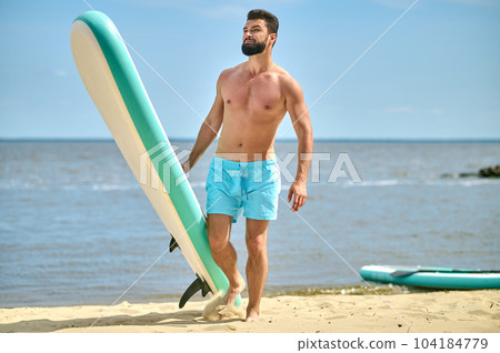 Handsome athletic man standing with paddle board on sandy beach near sea, enjoying vacation. Handsome athletic man standing with paddle board on sandy beach near sea, enjoying vacation. 104184779