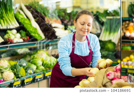 Skillful busy middle-aged female seller in uniform putting fresh onion on shelves in grocery shop Skillful busy middle-aged female seller in uniform putting fresh onion on shelves in grocery shop 104186691