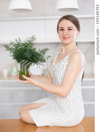 Woman in dress with short sleeves sitting near kitchen table and holding glass with bunch of dill 104186763