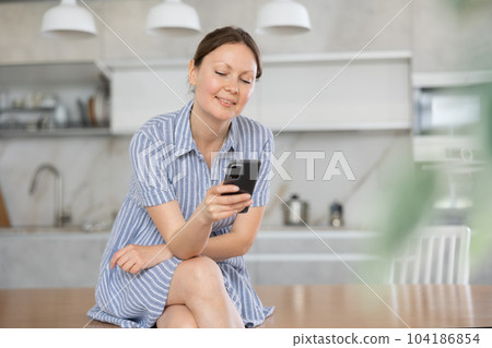 In kitchen, positive girl in casual clothes sits on table and chatting in smartphone 104186854