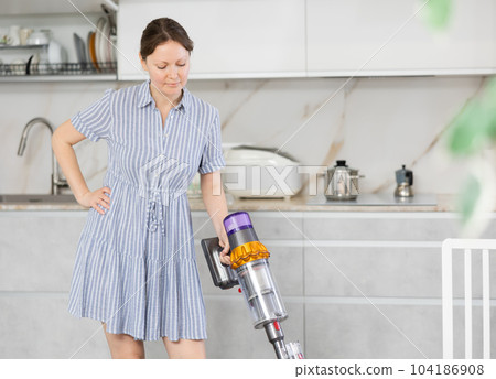 Smiling housewife in blue dress run vacuum cleaner floor covering near dining table 104186908