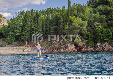 Young women Having Fun Stand Up Paddling in blue water sea near st stefan island against the backdrop of Milocer Park in Montenegro. SUP Young women Having Fun Stand Up Paddling in blue water sea near st stefan island against the backdrop of Milocer Park in Montenegro. SUP 104189261