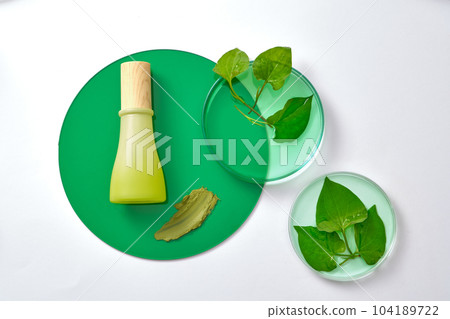 A green bottle unlabeled and fresh fish mint leaves on petri dish decorated with acrylic sheet on white background. Mockup for design, organic cosmetic of fish mint extract, beauty product concept 104189722