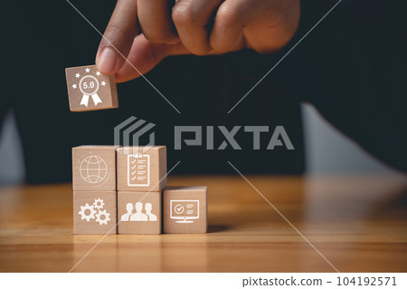 Quality control and assurance are represented by a hand placing wooden cubes with ISO icons on a table. An ISO banner in the background signifies certification and standardization 104192571