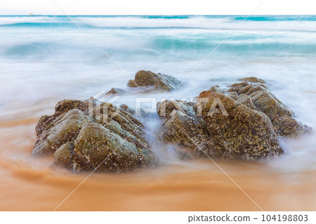 Dramatic moving wave around big rocks on clear water beach. Dramatic moving wave around big rocks on clear water beach. 104198803