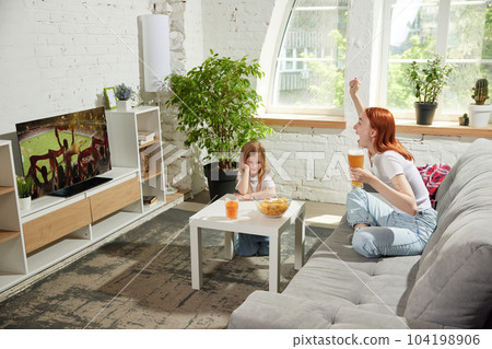 Young loud, excited woman sitting on couch at home, drinking beer and emotionally watching football match on tv. Bored child looking 104198906