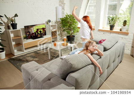 Young excited woman sitting on couch at home and emotionally watching football match on tv while little girl, child feeling bored 104198934