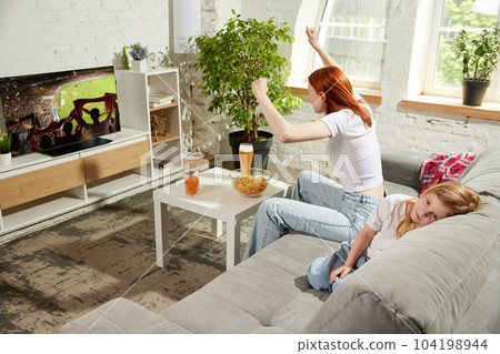 Young woman sitting on couch at home and attentively watching football match on tv. Little girl, child feeling bored and sleepy 104198944