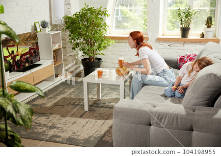 Young woman sitting on couch at home and attentively watching football match on tv. Little girl, child feeling bored and sleepy 104198955