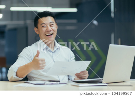 Portrait of a young Asian businessman man sitting at a desk in the office with a laptop and holding documents. rejoices in a successful deal and increased income. Pointing finger at camera super Portrait of a young Asian businessman man sitting at a desk in the office with a laptop and holding documents. rejoices in a successful deal and increased income. Pointing finger at camera super 104199567
