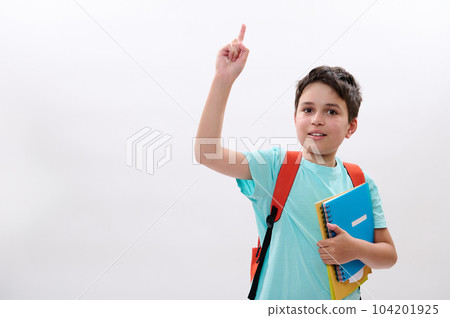 Smart schoolboy, handsome preteen boy with backpack and school supplies, expressing positive emotions, smiles looking at camera, pointing aside a copy space for your ads on white isolated background 104201925