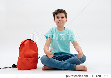 Handsome Caucasian teenager schoolboy in blue t-shirt and denim, sitting barefoot in lotus pose near his orange backpack, thumbing up, smiling, looking at camera, isolated over white studio background Handsome Caucasian teenager schoolboy in blue t-shirt and denim, sitting barefoot in lotus pose near his orange backpack, thumbing up, smiling, looking at camera, isolated over white studio background 104201986