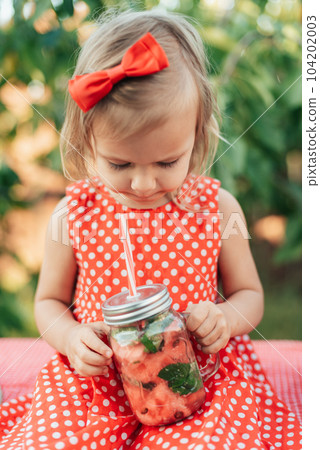 Lemonade Stand. Adorable little girl trying to sell lemonade. Watermelon lemonade with ice and mint as summer refreshing drink in jars. Cold soft drinks with fruit. Child drinking lemonade in jar Lemonade Stand. Adorable little girl trying to sell lemonade. Watermelon lemonade with ice and mint as summer refreshing drink in jars. Cold soft drinks with fruit. Child drinking lemonade in jar 104202003