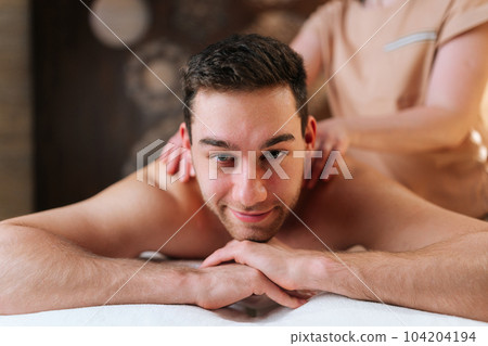 Closeup face of handsome bearded man getting relaxing massage and smiling looking at camera lying on massage table. Unrecognizable professional masseuse doing back revitalizing massage to client. Closeup face of handsome bearded man getting relaxing massage and smiling looking at camera lying on massage table. Unrecognizable professional masseuse doing back revitalizing massage to client. 104204194