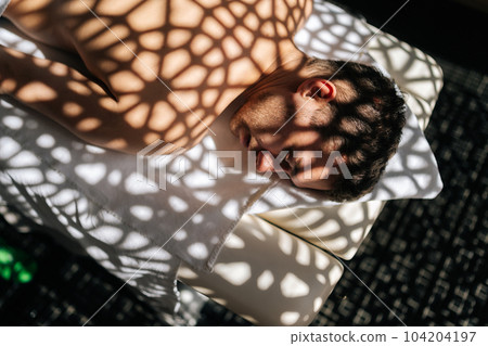 Close-up top view of handsome young man relaxing lying down with closed eyes on massage table in sunlight and beautiful shadows in spa salon. Concept of body care and rehabilitation of spiritual peace 104204197