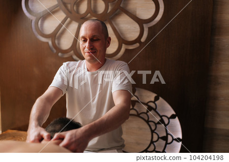 Portrait of positive professional male masseur massaging neck and back of unrecognizable sporty man lying on massage table in luxury spa salon. Concept of healthy lifestyle and body care. Portrait of positive professional male masseur massaging neck and back of unrecognizable sporty man lying on massage table in luxury spa salon. Concept of healthy lifestyle and body care. 104204198