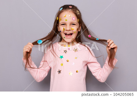 Indoor shot of cheerful joyful little brown haired little girl covered with stickers posing isolated over gray background, expressing happiness, pulling her hair aside. 104205065