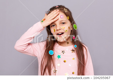 Indoor shot of funny little brown haired little girl covered with stickers posing isolated over gray background, forgot rules of game, showing facepalm gesture. Indoor shot of funny little brown haired little girl covered with stickers posing isolated over gray background, forgot rules of game, showing facepalm gesture. 104205067
