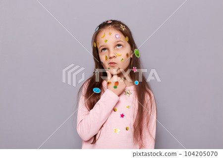 Photo of thoughtful pensive little brown haired little girl covered with stickers posing isolated over gray background, holding her chin, thinking, pondering. 104205070