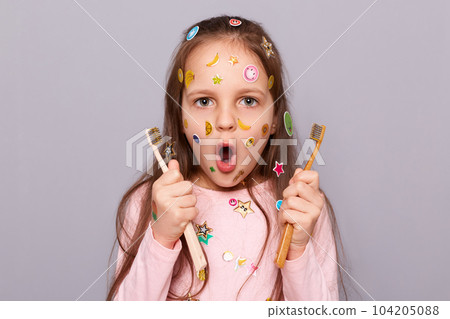 Shocked little girl covered with colorful stickers holding two toothbrushes, looking at camera with amazed look, being surprised, oral care, wearing pink shirt standing isolated over gray background. 104205088