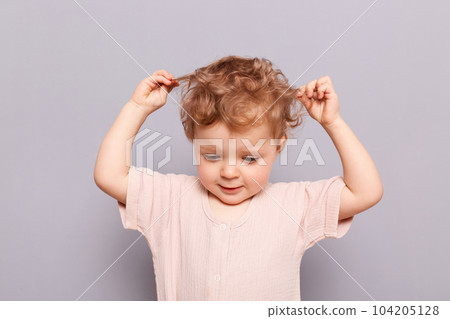 Indoor shot of beautiful adorable happy cute charming baby infant girl pulling her wavy blond hair, looking down, posing alone isolated over gray background. Indoor shot of beautiful adorable happy cute charming baby infant girl pulling her wavy blond hair, looking down, posing alone isolated over gray background. 104205128