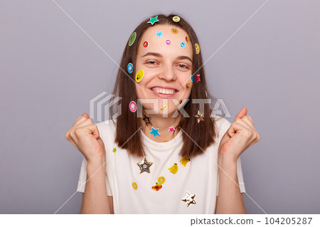 Indoor shot of happy cheerful woman covered with funny stickers clenched fists, celebrating her success posing isolated over gray background, smiling toothily. 104205287