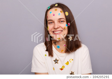 Indoor shot of pretty attractive smiling woman covered with funny stickers posing isolated over gray background, looking at camera with charming smile, rejoicing. 104205291