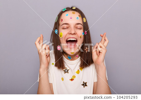 Horizontal shot of extremely happy woman covered with funny stickers posing isolated over gray background, crossing her fingers,closed eyes, praying for good luck and success. Horizontal shot of extremely happy woman covered with funny stickers posing isolated over gray background, crossing her fingers,closed eyes, praying for good luck and success. 104205293