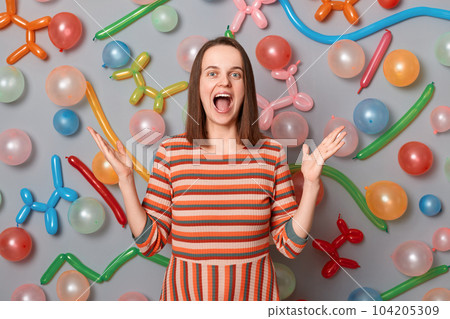 Indoor shot of extremely happy joyful woman with brown hair wearing striped dress posing over gray background with inflated balloons, celebrating birthday, having pleasant surprise Indoor shot of extremely happy joyful woman with brown hair wearing striped dress posing over gray background with inflated balloons, celebrating birthday, having pleasant surprise 104205309