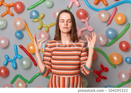 Indoor shot of calm relaxed woman with brown hair wearing striped dress posing isolated over gray background with different shaped balloons, keeps hands in mudra gesture, meditating. 104205322