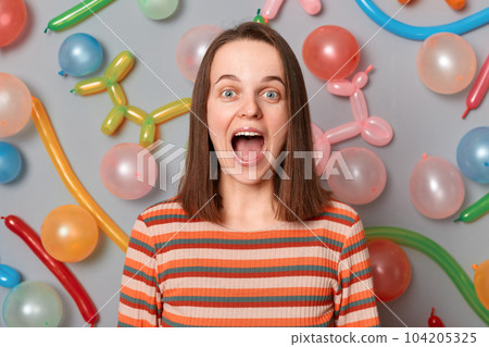 Extremely happy excited woman with brown hair wearing striped dress, looking at camera with big eyes, screaming with happiness, posing isolated over gray background with different shaped balloons 104205325