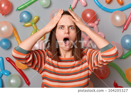 Portrait of astonished shocked scared woman with brown hair wearing striped dress posing isolated over gray background with different shaped balloons, keeps hands on head, screaming with surprise. 104205327