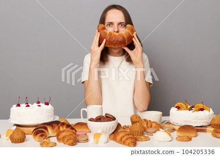 Indoor shot of funny serious woman with brown hair sitting at table isolated over gray background, having dinner, posing among sweet pastry, covering mouth with bagel, having fun. 104205336