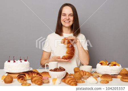 Portrait of smiling satisfied cheerful woman with brown hair sitting at table isolated over gray background, showing homemade bagel, looking at camera with toothy smile, advertising bakery. 104205337