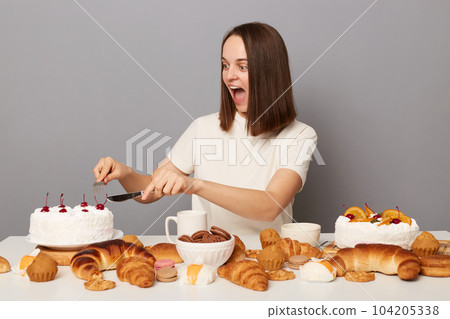 Photo of amazed surprised excited woman with brown hair sitting at table isolated over gray background, holding fork and knife and eating white delicious cake. 104205338