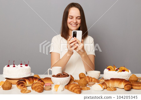 Delighted smiling cheerful millennial woman with brown hair wearing white T-shirt sitting at table chatting online via cell phone, enjoys eating desserts, isolated over gray background 104205345