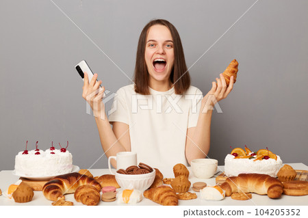 Amazed woman with brown hair sitting at table with mobile phone in hand among various desserts, has good appetite excited with variety of confectionery, tired of diets isolated over gray background 104205352