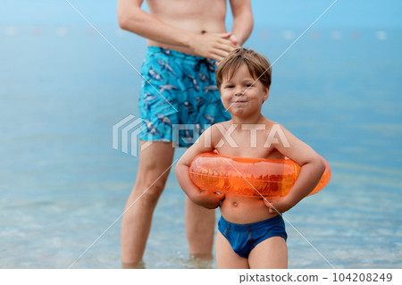 Son and father are standing on the beach after swimming in the sea. The child has an inflatable circle. Son and father are standing on the beach after swimming in the sea. The child has an inflatable circle. 104208249