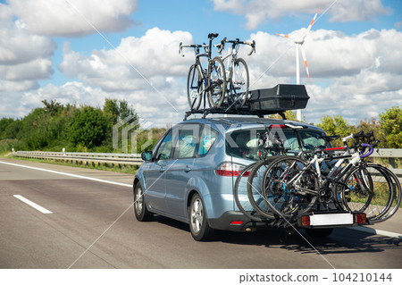 Back view modern white family wagon car with mounted roof box trunk and bike tail carrier driving european highway road against blue sky summer day. Lifestyle travel adventure trip journey concept Back view modern white family wagon car with mounted roof box trunk and bike tail carrier driving european highway road against blue sky summer day. Lifestyle travel adventure trip journey concept 104210144