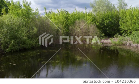 the drone flies over the small lake and takes off above the trees. Green nature with mountain views. Aerial photography. 104210366