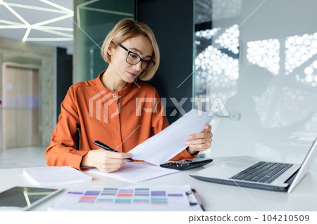 Serious and pensive business woman behind paper work inside office, female financier worker thinks about contracts and reports with charts and graphs, blonde successful woman uses laptop at work. 104210509