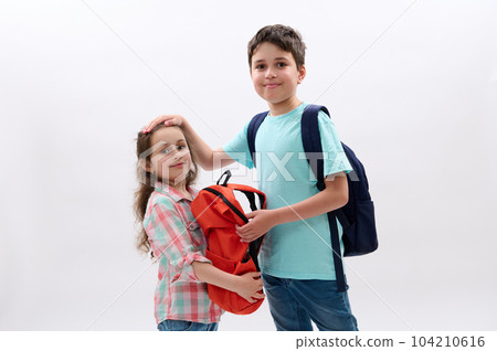 Caucasian handsome preteen child boy with backpack, strokes his younger sister, ready to go at school, starting new semester of academic year, smiling looking at camera, isolated over white background 104210616