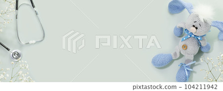 Kids health care banner. Cute knitted toy rabbit with a stethoscope on blue background with white flowers. Family doctor or pediatrician concept. Banner size. Copy space for text 104211942
