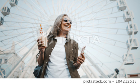 Happy mature woman smiling against urban city background. She seen enjoying refreshing drink from cup while also engaging with mobile phone, possibly texting message. . High quality photo Happy mature woman smiling against urban city background. She seen enjoying refreshing drink from cup while also engaging with mobile phone, possibly texting message. . High quality photo 104212515