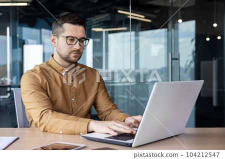Portrait of a serious young male programmer, specialist developer who is concentrating on working on a laptop while sitting at a desk in an office center. 104212547