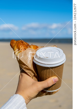 Tasty fresh croissants with coffee in paper cup in female hands on beach sea background. Breakfast outdoors wellbeing concept. FPV. Take away food 104212677