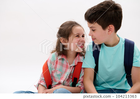Mischievous little girl, shows tongue and makes faces, smiles to her older brother, isolated over white background. Cheerful smart school Kids with backpacks having fun together. Family relationships 104213363