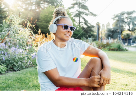 Smiling mixed race young woman with a rainbow badge, symbol of LGBTQ social community listening music with wireless headphones while relax in park. Social Diversity acceptance, feel free lifestyle. Smiling mixed race young woman with a rainbow badge, symbol of LGBTQ social community listening music with wireless headphones while relax in park. Social Diversity acceptance, feel free lifestyle. 104214116