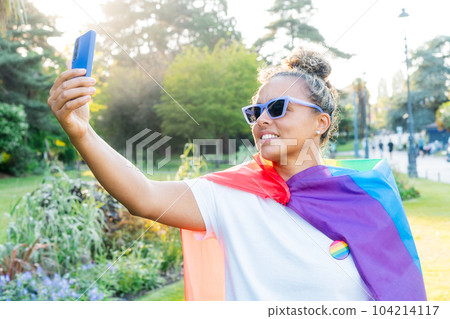 Young Latin happy woman wrapped in a rainbow flag, symbol of LGBT community, makes selfie on a mobile phone on a pride celebration in a city park. Human Rights, Equality, LGBT, Freedom and Happiness Young Latin happy woman wrapped in a rainbow flag, symbol of LGBT community, makes selfie on a mobile phone on a pride celebration in a city park. Human Rights, Equality, LGBT, Freedom and Happiness 104214117